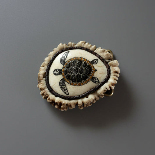 A circular handmade elk antler belt buckle featuring a sea turtle design, displayed on a gray surface.