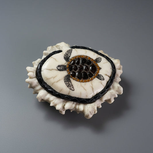 A handmade turtle-shaped belt buckle, made of moose antler cornet featuring a cream-colored textured shell, a black braided border, and a central turtle design with black, brown, and gold details, set against a gray background.